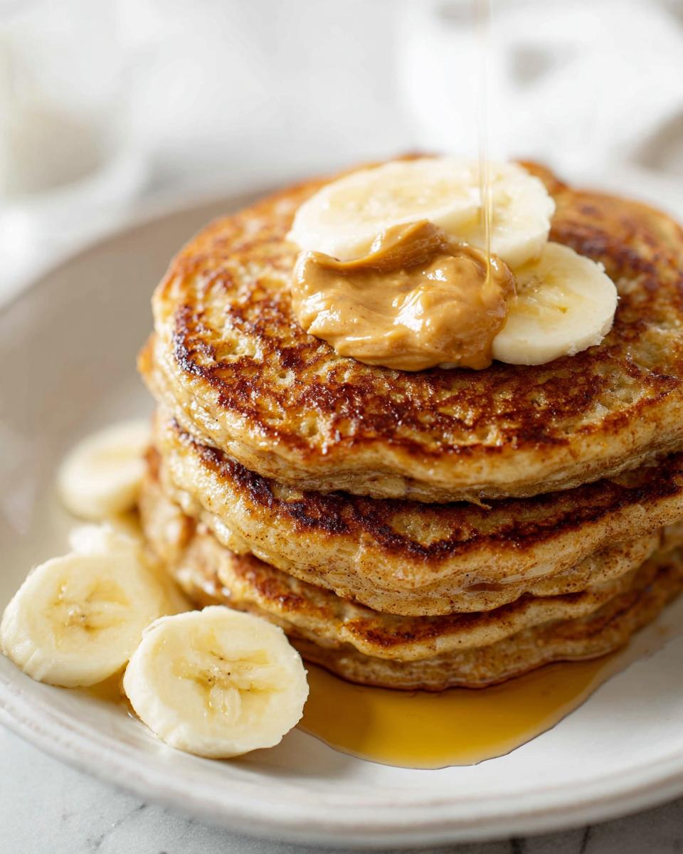 A stack of golden-brown Banana Oat Pancakes topped with peanut butter, banana slices, and syrup.