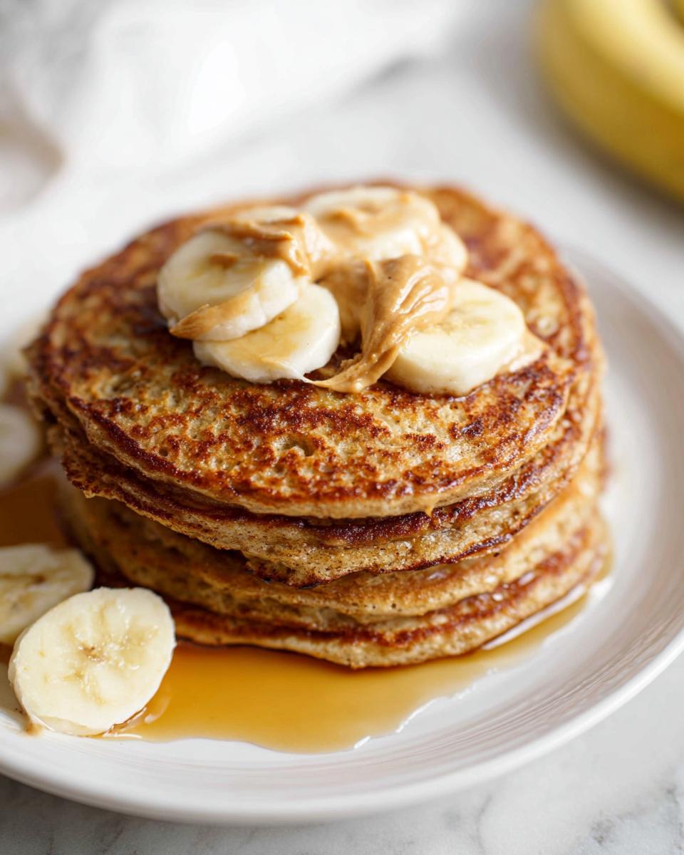 A tall stack of golden Banana Oat Pancakes topped with sliced bananas and a dollop of peanut butter, drizzled with syrup.