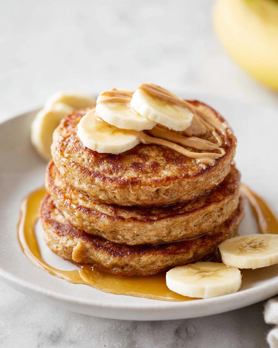 A stack of three golden Banana Oat Pancakes topped with peanut butter and banana slices, drizzled with syrup.