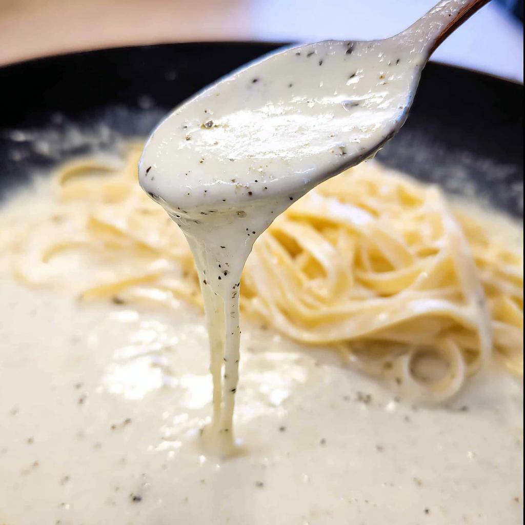 A close-up of a spoon lifting creamy Alfredo sauce with black pepper over a plate of fettuccine pasta.