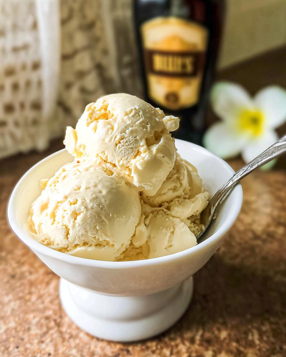 A white bowl filled with creamy scoops of homemade Baileys ice cream, with a blurred bottle of Baileys and a flower in the background.
