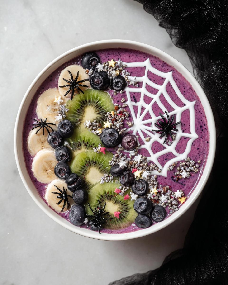 A vibrant purple Spooky Halloween Smoothie Bowl topped with kiwi, blueberries, bananas, and a spiderweb design with candy spiders.