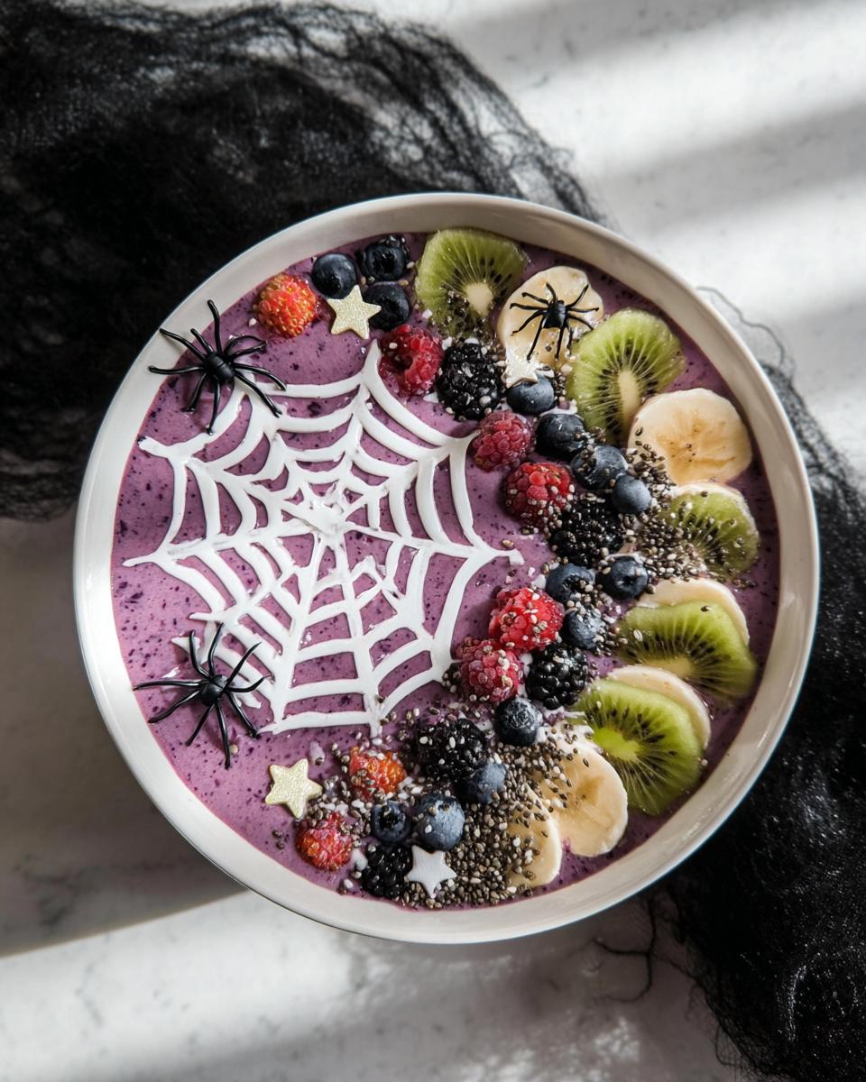 A Spooky Halloween Smoothie Bowl decorated with a white spiderweb, berries, kiwi, banana slices, and plastic spiders.
