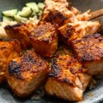 Close-up of crispy-edged Air Fryer Salmon cubes served over brown rice with sliced cucumbers.