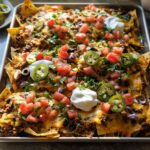 A large sheet pan filled with fully loaded Sheet Pan Nachos, topped with melted cheese, ground meat, tomatoes, jalapeños, and sour cream.