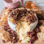 A wheel of Baked Brie with Honey and Pecans, melted and oozing, served with toasted bread slices on a wooden board.