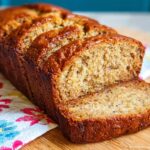 A freshly baked loaf of Banana Bread with Sour Cream, partially sliced on a wooden board.