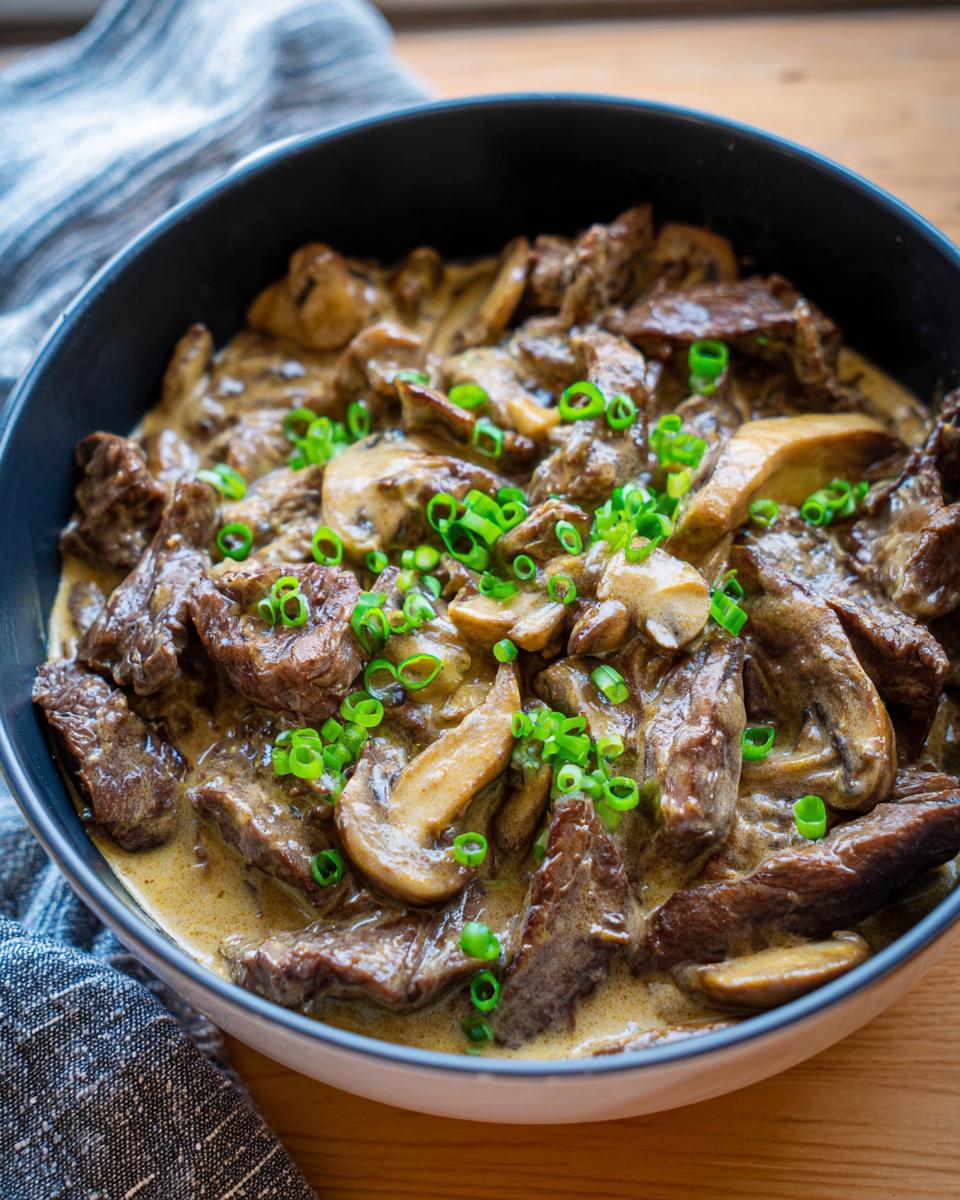 A close-up of a bowl filled with creamy Beef Stroganoff, featuring tender beef strips, sautéed mushrooms, and fresh green scallions.
