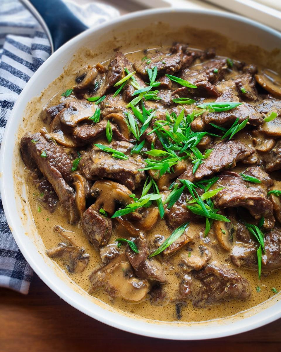 A close-up view of a skillet filled with creamy Beef Stroganoff, featuring tender beef strips and mushrooms, garnished with fresh green onions.