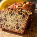 Close-up of moist Best Ever Banana Bread loaf sliced on a wooden board with a banana blurred in the background.