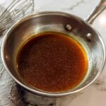 Close-up of rich, brown homemade au jus recipe simmering in a small stainless steel saucepan.