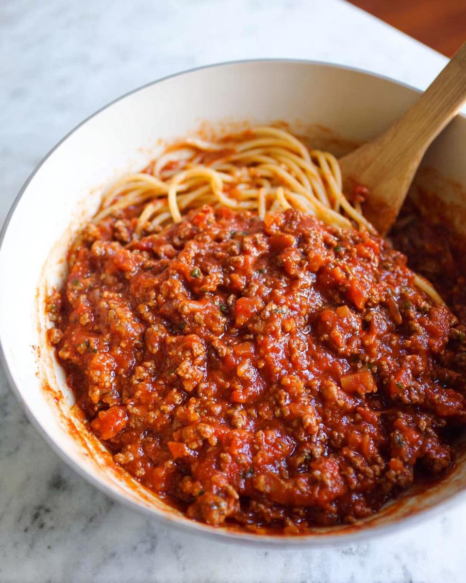 A close-up of a bowl filled with Classic Spaghetti with Meat Sauce, with a wooden spoon resting inside.