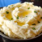 A close-up of a bowl filled with fluffy Creamy Mashed Potatoes, topped with melted butter and fresh parsley.