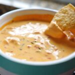 A hand dips a crispy tortilla chip into a bowl of creamy, orange Queso Dip containing visible pieces of tomato and cilantro.
