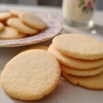 Close-up of perfectly baked, plain round Cutout Sugar Cookies with sharp edges, stacked next to a plate of more cookies and a glass of milk.