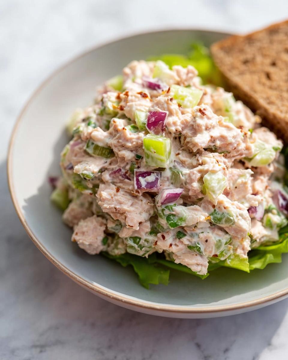 A close-up of a bowl of easy tuna salad meal prep, featuring flaked tuna mixed with chopped celery, red onion, and a creamy dressing, served on lettuce with a slice of toast.