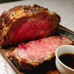 A perfectly cooked Garlic-Herb Prime Rib, sliced to reveal a medium-rare center, served with au jus on a wooden board.