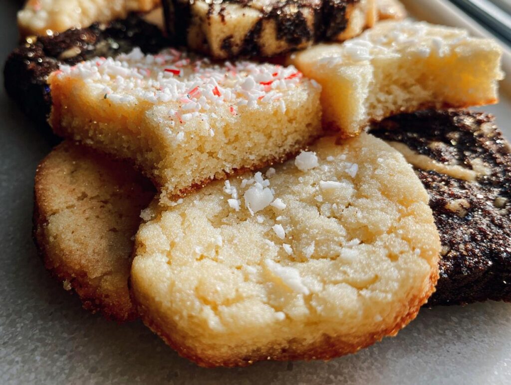 Close-up of various Gold-Dusted Shortbread cookies, some topped with sugar or crushed peppermint.