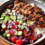 A close-up of a Greek Chicken Salad Bowls featuring grilled sliced chicken over rice, topped with cucumber, tomato, red onion, olives, and feta.