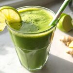A glass of vibrant Green Detox Smoothie garnished with a lime slice and a striped straw, next to fresh ginger roots.