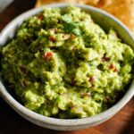 Close-up of chunky Guacamole with Lime and Cilantro served in a rustic bowl with tortilla chips nearby.