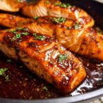 Close-up of glistening Honey Garlic Salmon fillets cooking in a dark skillet with a rich, thick glaze and fresh parsley.