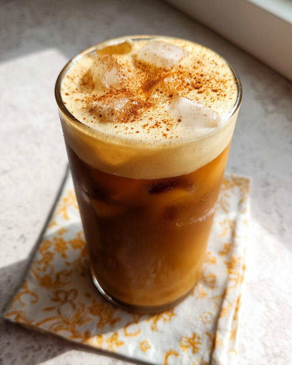 A tall glass of iced Pumpkin Cream Cold Brew topped with thick foam and a dusting of cinnamon spice.