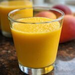 Close-up of a vibrant, thick Mango Pineapple Smoothie served in a clear glass, with whole mangoes blurred in the background.