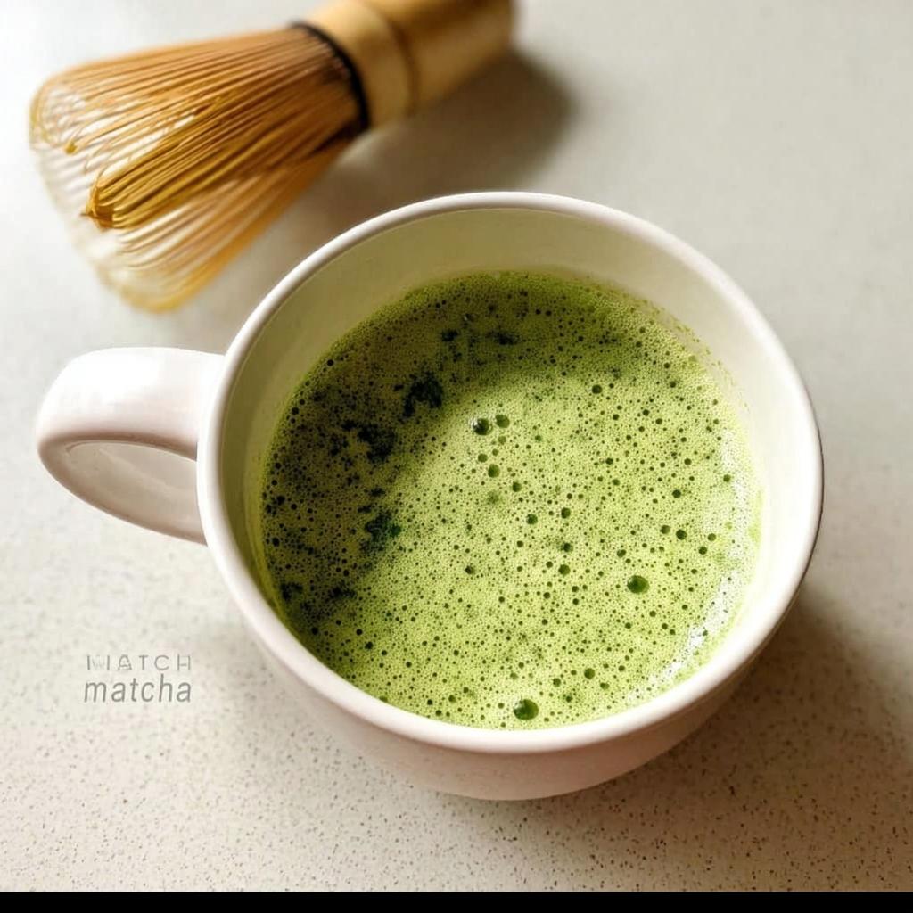 A freshly prepared Matcha Green Tea Latte with vibrant green foam in a white mug, with a bamboo whisk visible in the background.