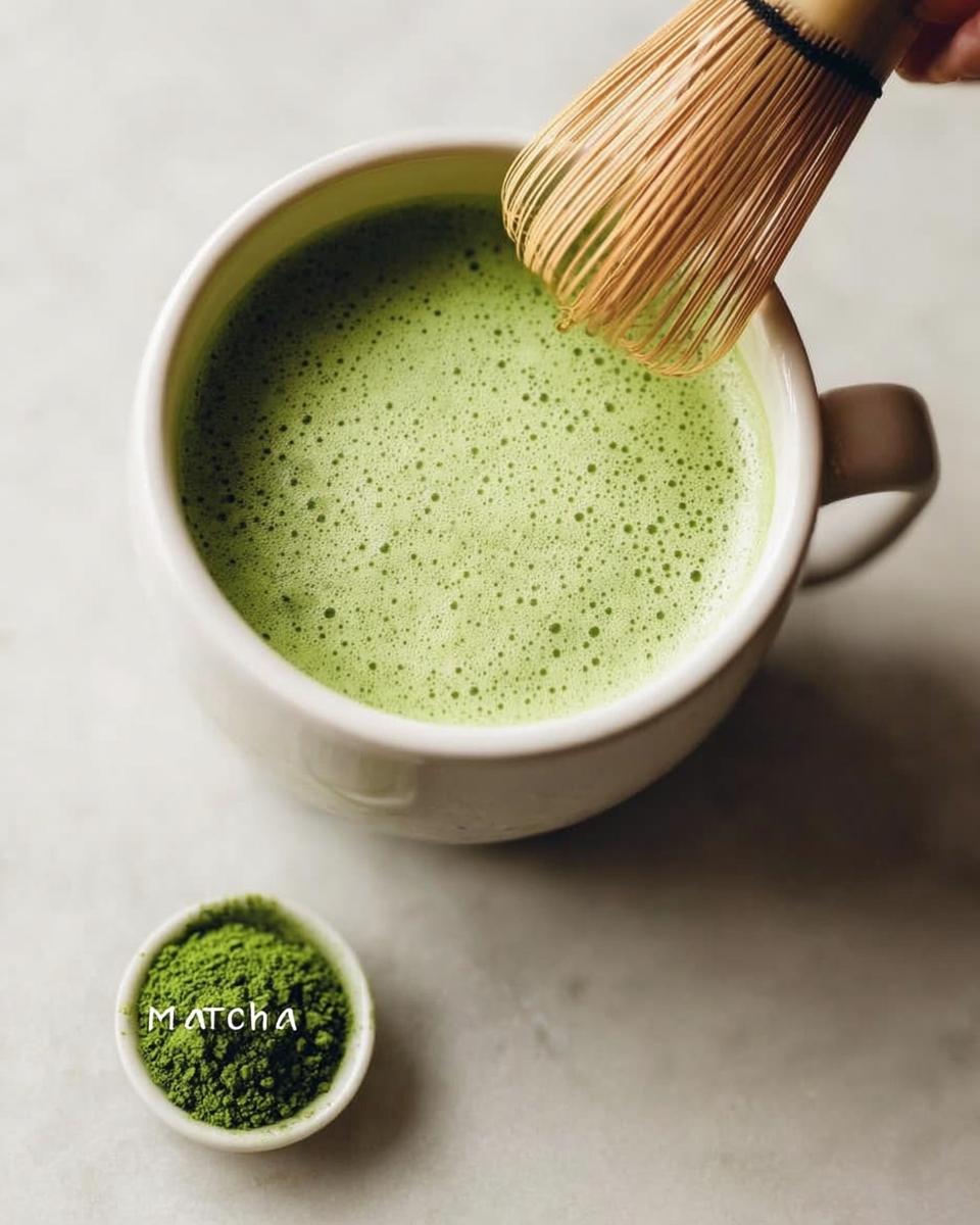 A bamboo whisk frothing a bright green Matcha Green Tea Latte in a white mug, with matcha powder below.