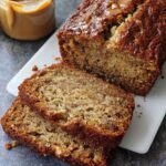 A loaf of rich Peanut Butter Banana Bread, partially sliced, drizzled with caramel sauce.