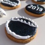 Close-up of round sugar cookies decorated for New Year’s Eve, featuring dark blue edible glitter icing and white piped borders. One cookie reads '2019'.