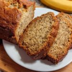 A freshly baked loaf of moist One Bowl Banana Bread, partially sliced on a white plate.