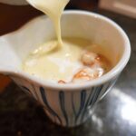 Pouring rich Champagne Cream Sauce over cooked shrimp in a decorative white and blue serving dish.