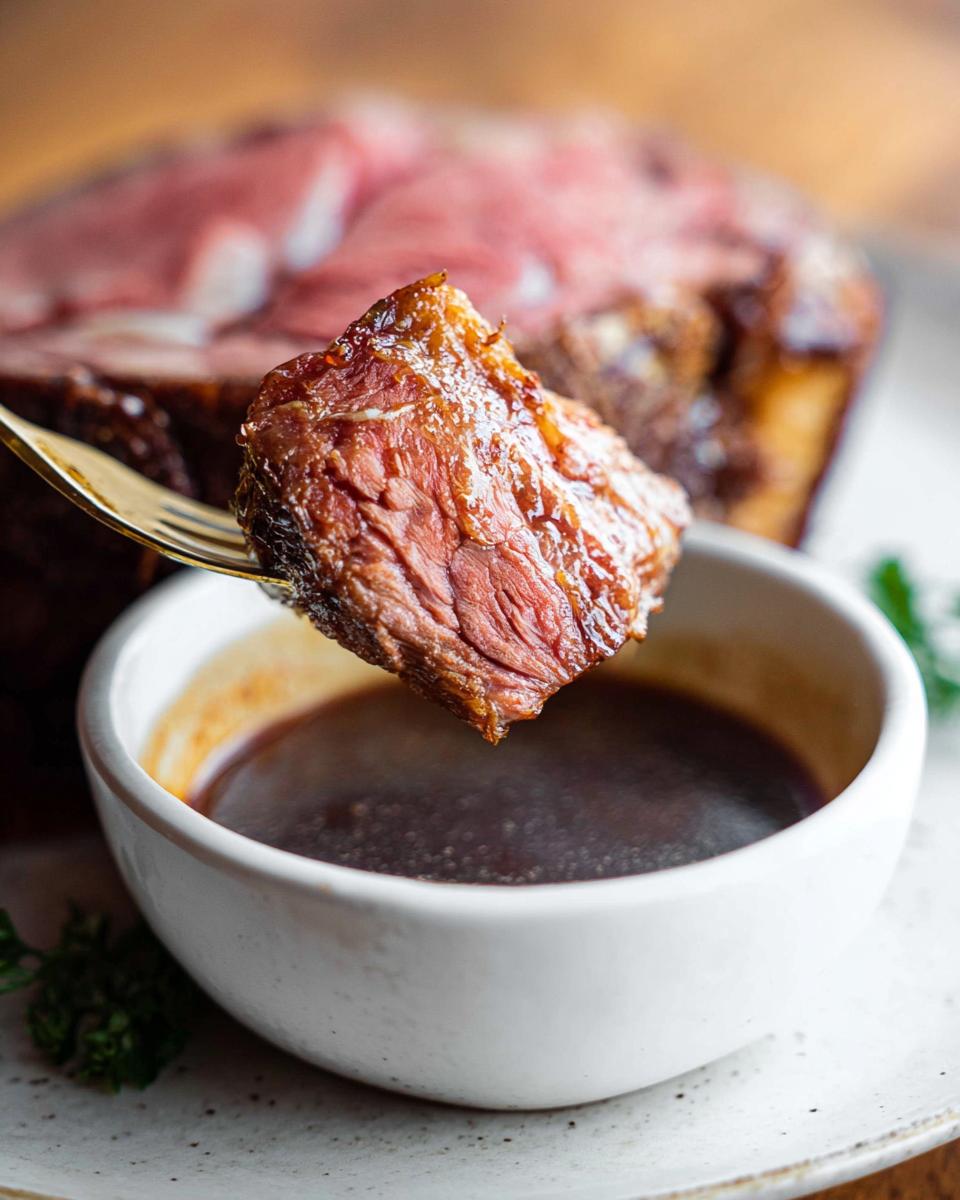 A close-up of a perfectly cooked piece of prime rib being dipped into a small bowl of rich, dark Prime Rib Au Jus Recipe sauce.