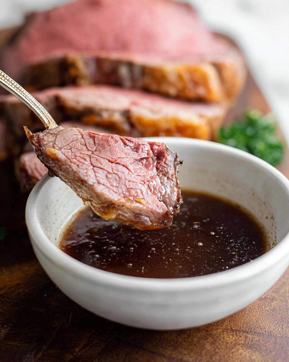 A slice of medium-rare prime rib being dipped into a small white bowl of rich Prime Rib Au Jus Recipe sauce.