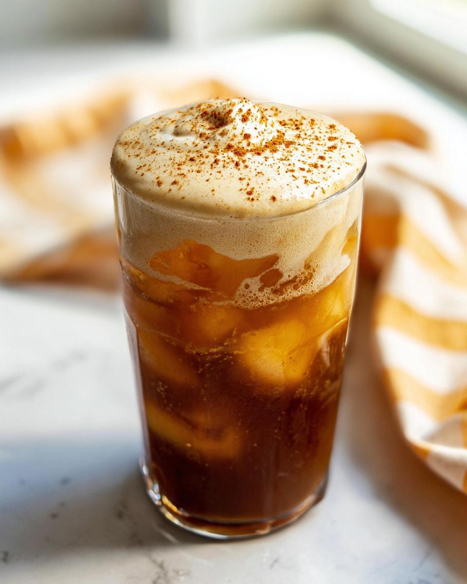 A tall glass filled with iced coffee and topped with thick, foamy Pumpkin Cream Cold Brew, dusted with spice.