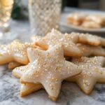 A stack of star-shaped Champagne Sugar Cookies topped with pale icing and gold/silver sprinkles.