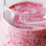 Close-up of a frothy pink Strawberry Oatmeal Smoothie with white swirls on top in a clear glass.