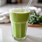 A tall glass filled with a vibrant Tropical Green Smoothie (Kale & Spinach) on a white countertop.