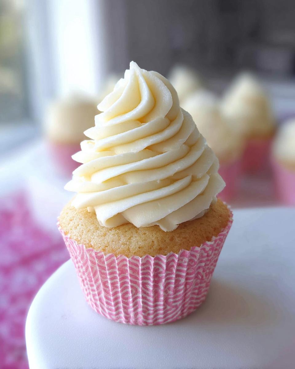 A single delicious vanilla cupcake topped with a generous swirl of creamy buttercream frosting in a pink patterned liner.