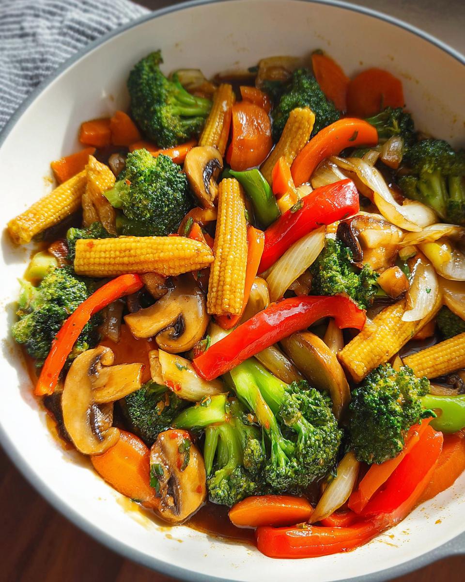 A close-up overhead view of a vibrant Vegetable Stir Fry in a white bowl, featuring broccoli, baby corn, mushrooms, carrots, and red bell peppers.