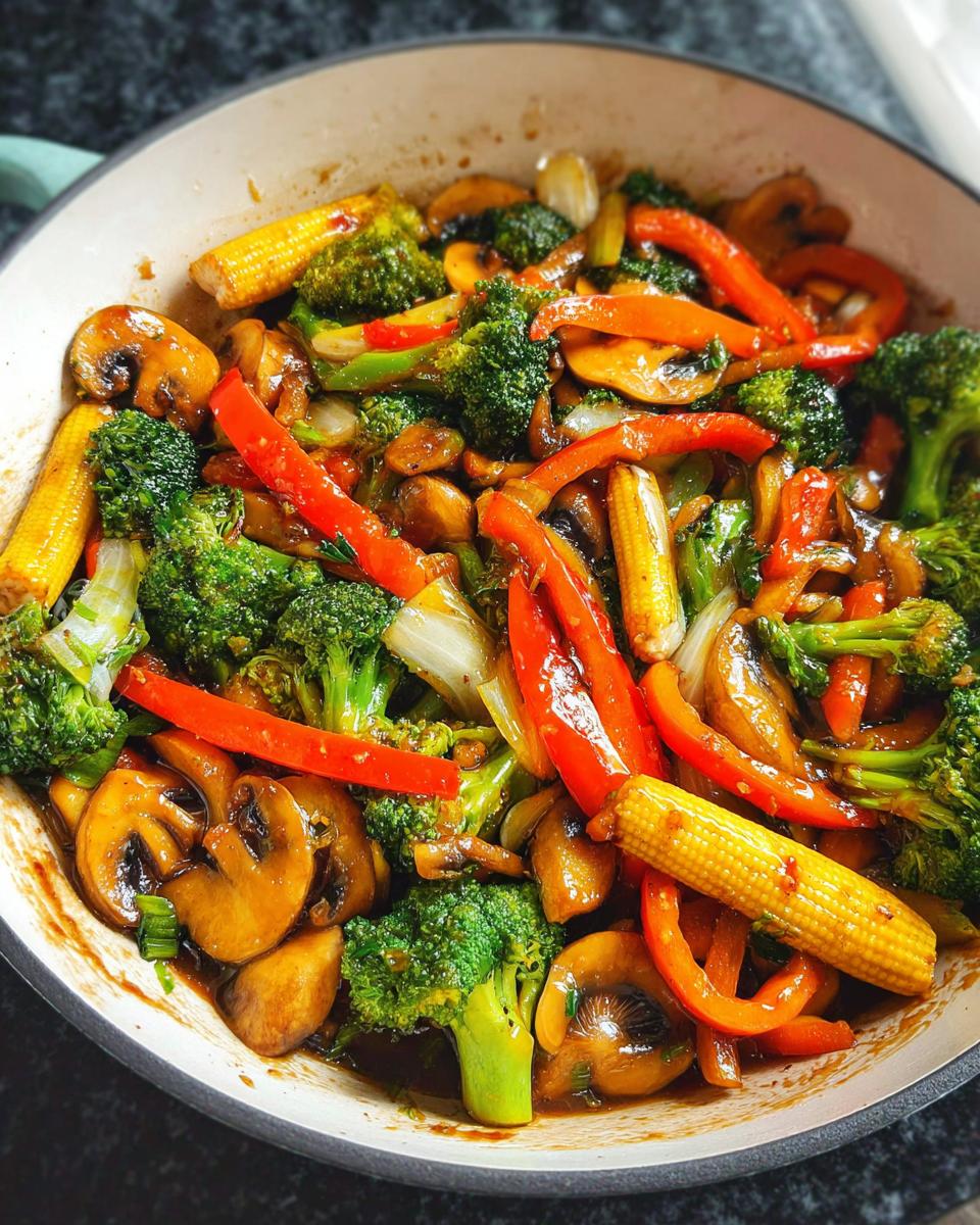 A close-up overhead view of a vibrant Vegetable Stir Fry in a pan, featuring broccoli, baby corn, mushrooms, and red bell peppers.
