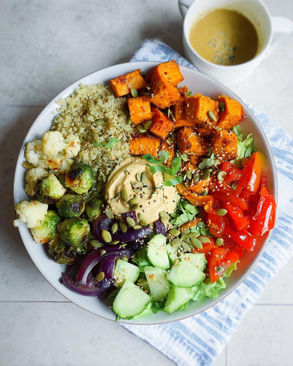 A vibrant and healthy vegetarian meal prep bowl filled with quinoa, roasted sweet potatoes, Brussels sprouts, cauliflower, red onion, cucumber, and hummus.