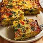 A square slice of Veggie Breakfast Casserole featuring layers of potatoes, spinach, and red peppers, being lifted from the main dish.