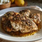 Four pieces of moist Baked Greek Lemon Oregano Chicken seasoned heavily with herbs on a white plate.