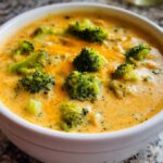 Close-up of a white bowl filled with rich, orange Creamy Broccoli Cheddar Soup topped with bright green broccoli florets.