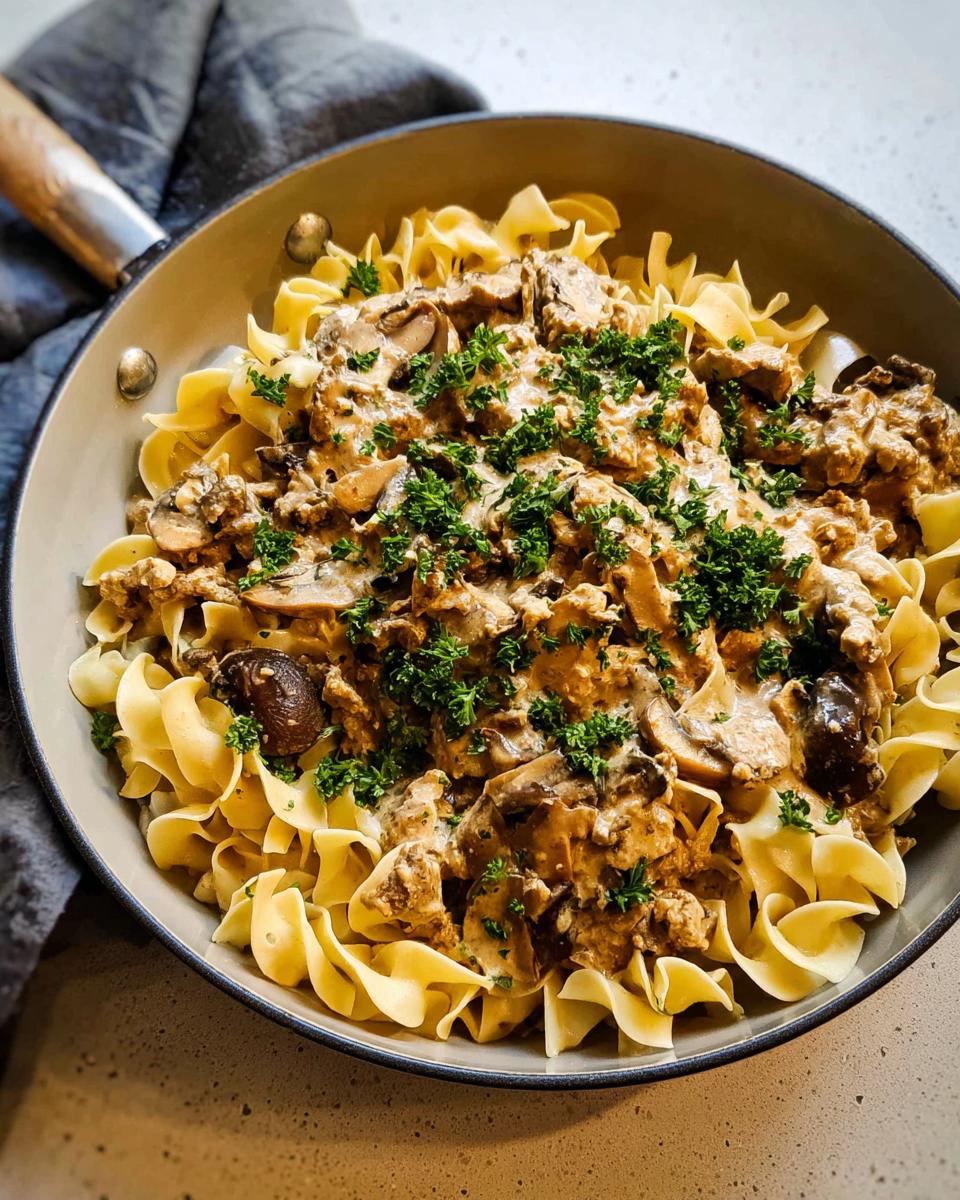 A skillet filled with Creamy Turkey Stroganoff sauce, mushrooms, and ground turkey served over wide egg noodles, garnished with parsley.
