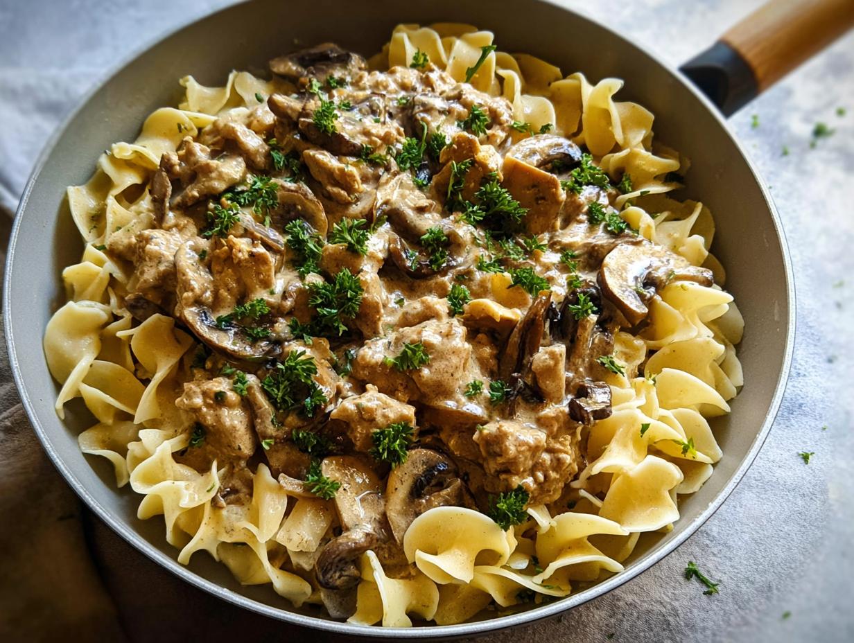 A skillet filled with Creamy Turkey Stroganoff sauce, mushrooms, and turkey pieces served over wide egg noodles, garnished with parsley.