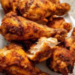 A pile of golden brown, seasoned Crispy Oven Baked Chicken Drumsticks served on a white platter, one piece broken open showing juicy white meat.